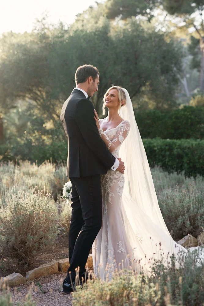 A bride and groom holding each other and looking into each other's eyes outdoors in a natural setting with trees and bushes, during sunset.