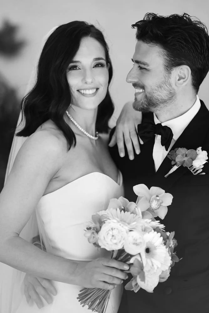 Black and white photo of a smiling bride and groom at their wedding, the bride holding a bouquet of flowers and wearing a strapless dress with a pearl necklace, and the groom in a tuxedo with a bow tie and boutonniere.