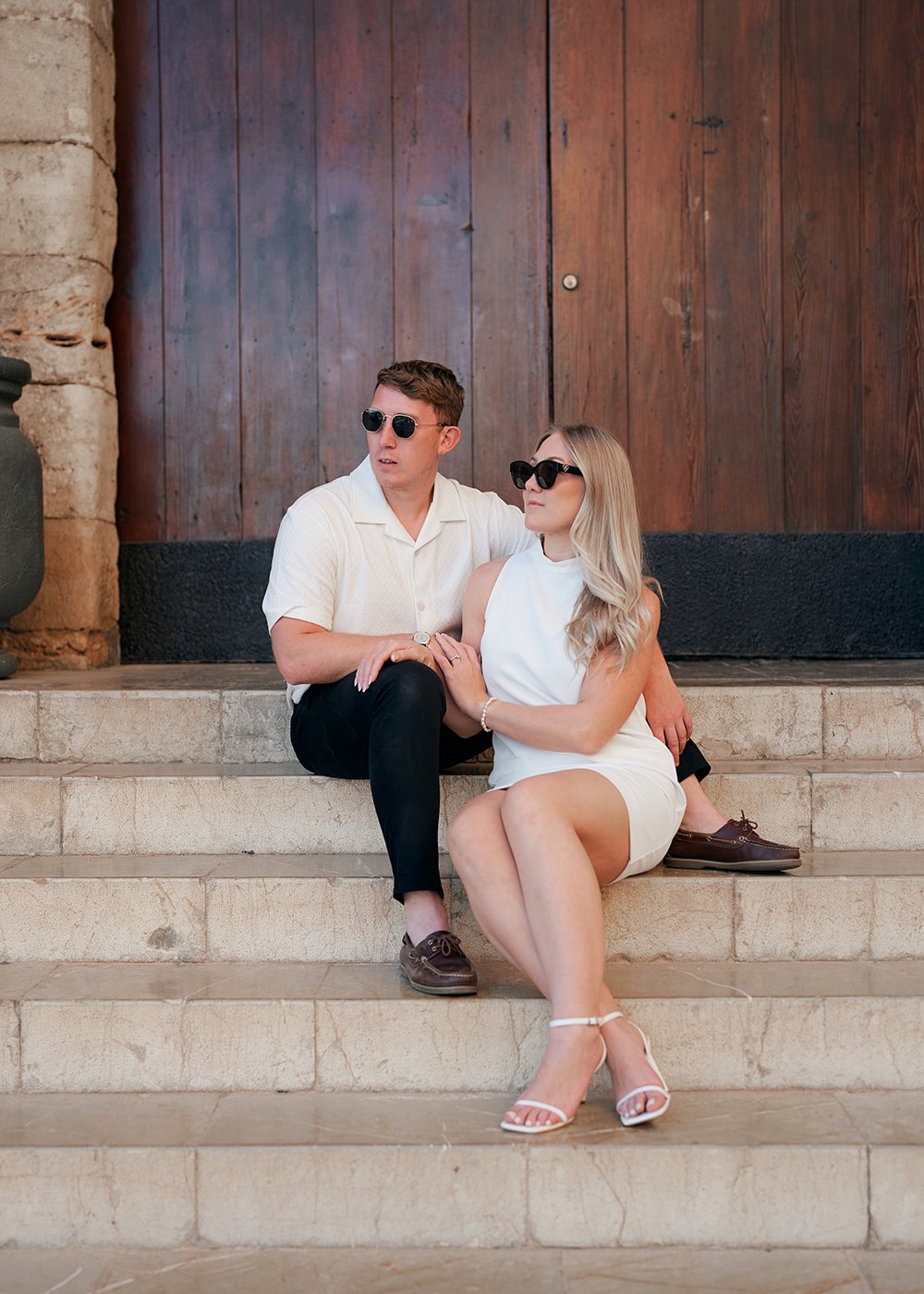 A couple sitting on stone steps wearing casual attire and sunglasses in front of a wooden door.