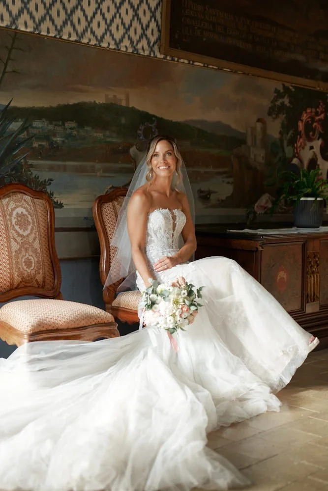 Bride in a white wedding dress with a long train, holding a bouquet, and sitting on vintage chairs in a decorated room with a mural and plants.