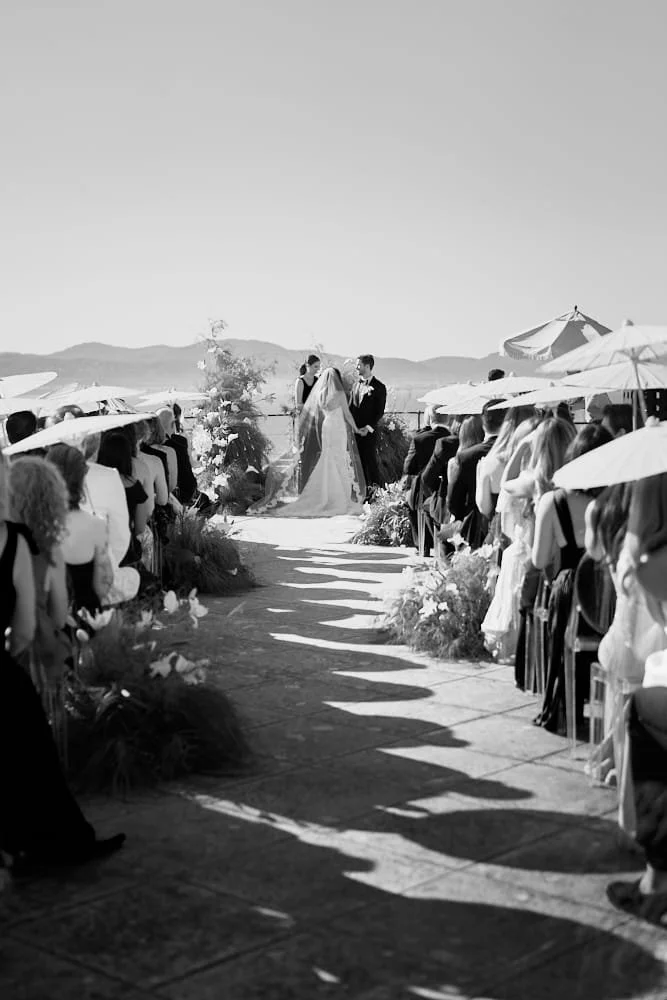 A black and white photo of a wedding ceremony outdoors with a bride and groom holding hands at the altar, surrounded by guests seated under umbrellas on both sides of a stone pathway.