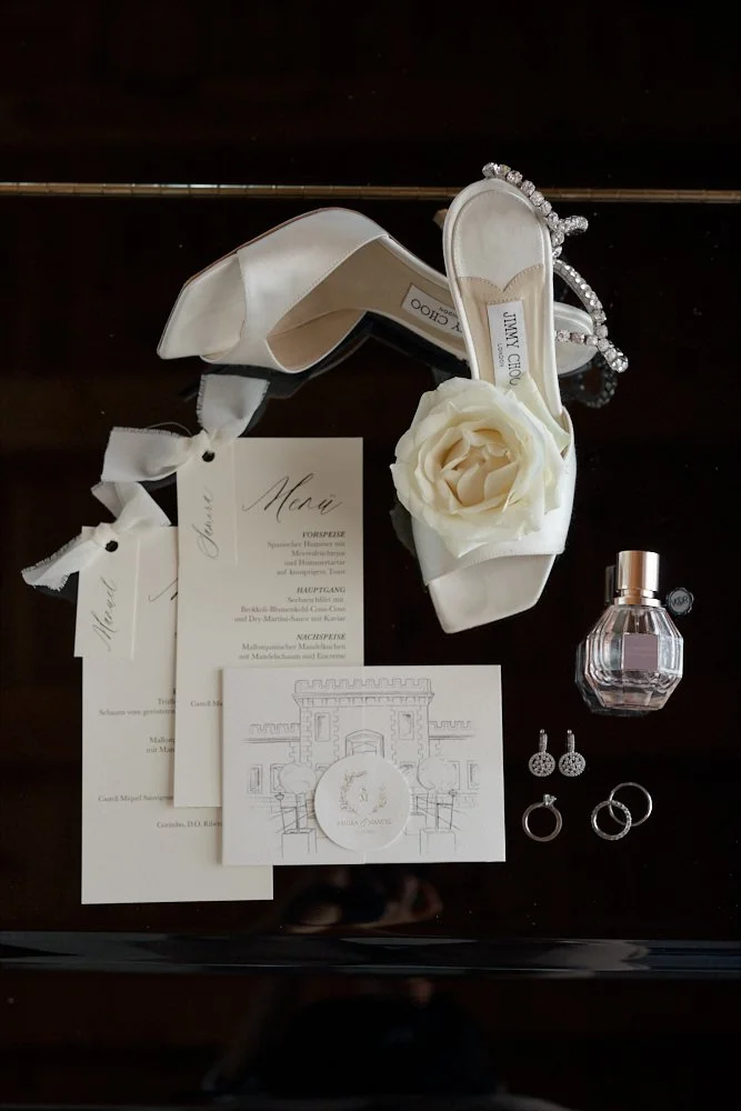 Wedding accessories including white high-heeled shoes with a large flower decoration, earrings, rings, a perfume bottle, and wedding programs on a dark surface.