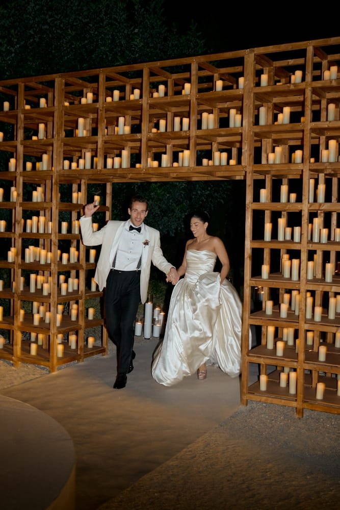 A bride and groom walking through a candle-lit archway at night, holding hands. The archway has a wooden grid structure filled with lit candles, creating a romantic atmosphere.