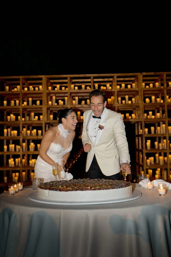 A bride and groom at their wedding reception, smiling and celebrating in front of a large cake on a table, with a backdrop of candles on wooden shelves.