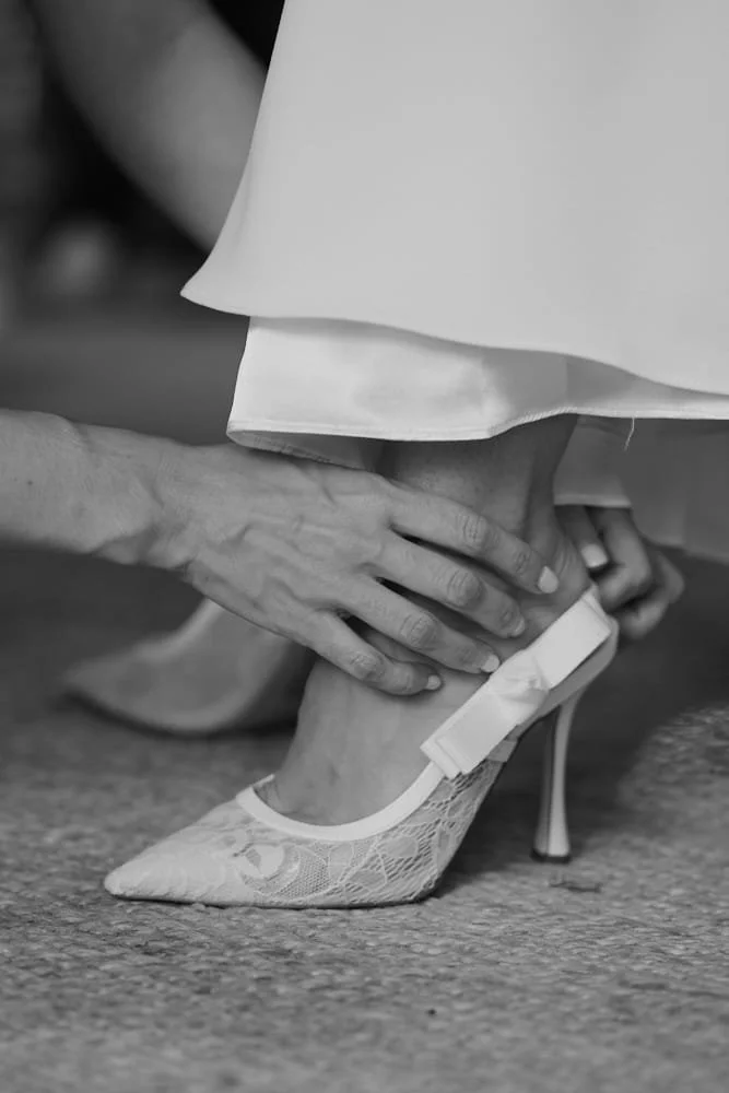 A person helping another put on a lace high-heeled shoe with a bow on the back, on a carpeted floor.