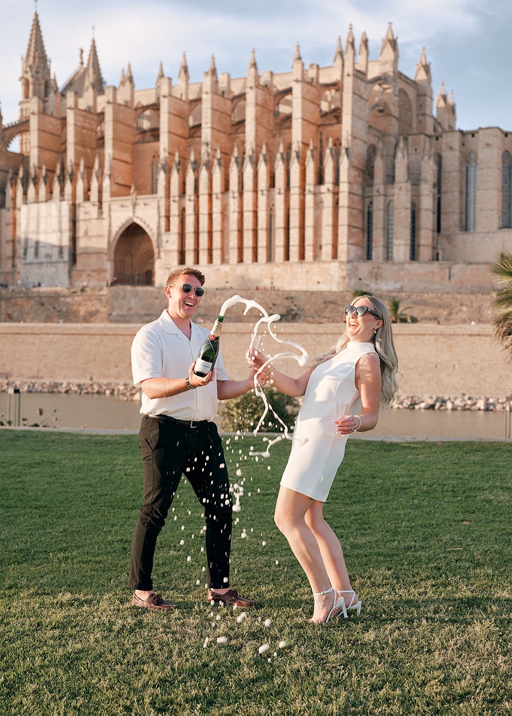 A man and woman celebrating with a champagne bottle in front of a cathedral in Palma de Mallorca, with champagne spraying and splashing around them. They are dressed stylishly and smiling, standing on green grass with the cathedral in the background.