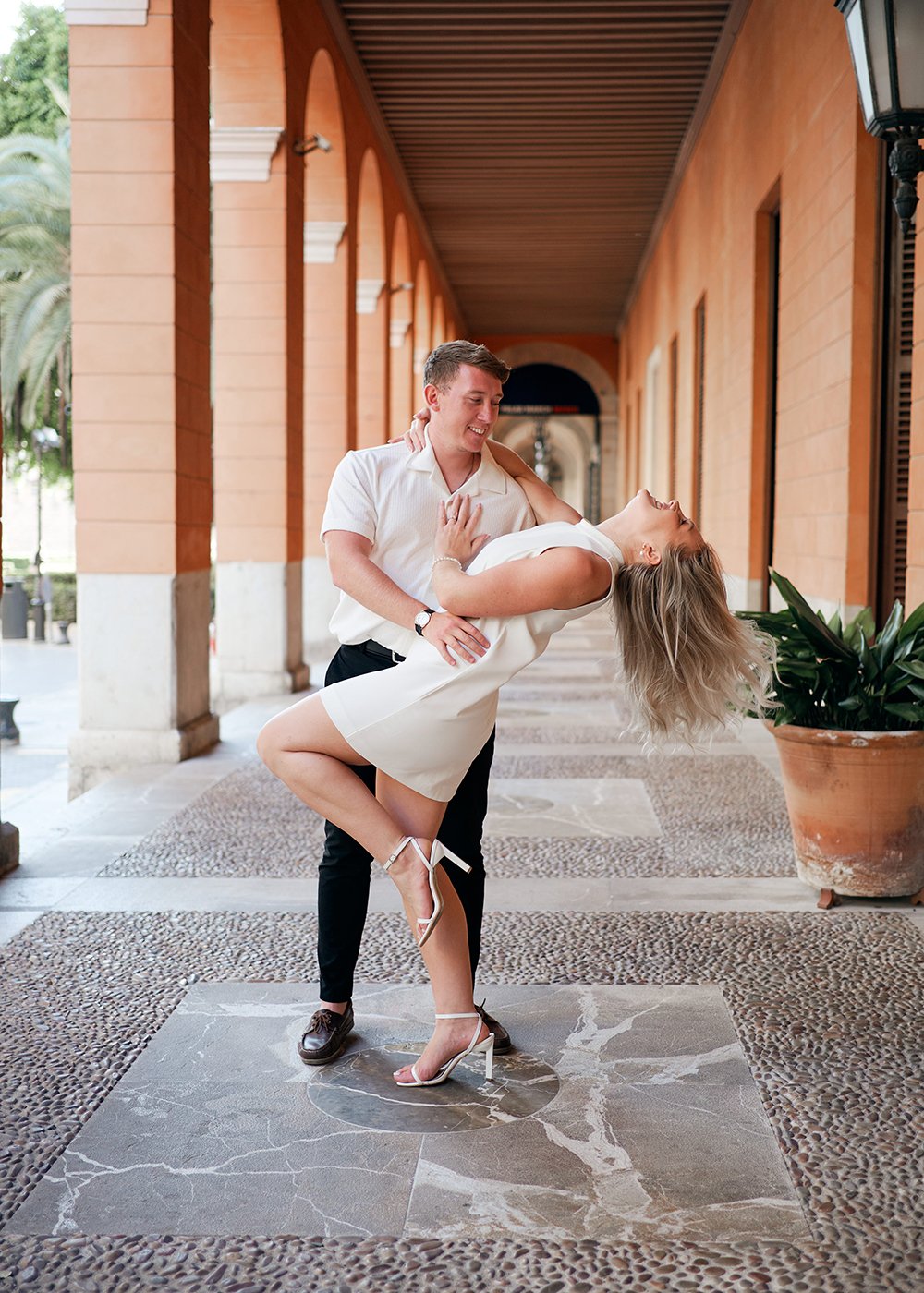 A man and woman dancing in an arched corridor, with the man dipping the woman. She is smiling and wearing a white dress and heels, while he is in a white shirt and dark pants. There are potted plants and decorative columns in the background.