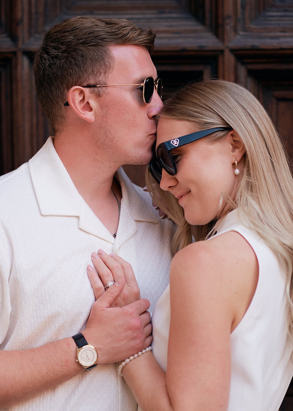 A man wearing sunglasses kisses a woman's forehead; both are smiling and wearing white outfits. The woman also wears sunglasses and an engagement ring, holding onto the man's chest.