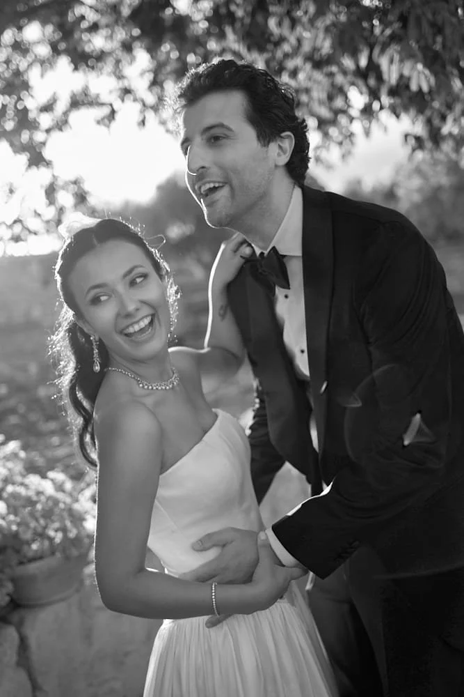 Black and white photo of a happy couple outdoors, dressed in formal attire, smiling and holding hands.