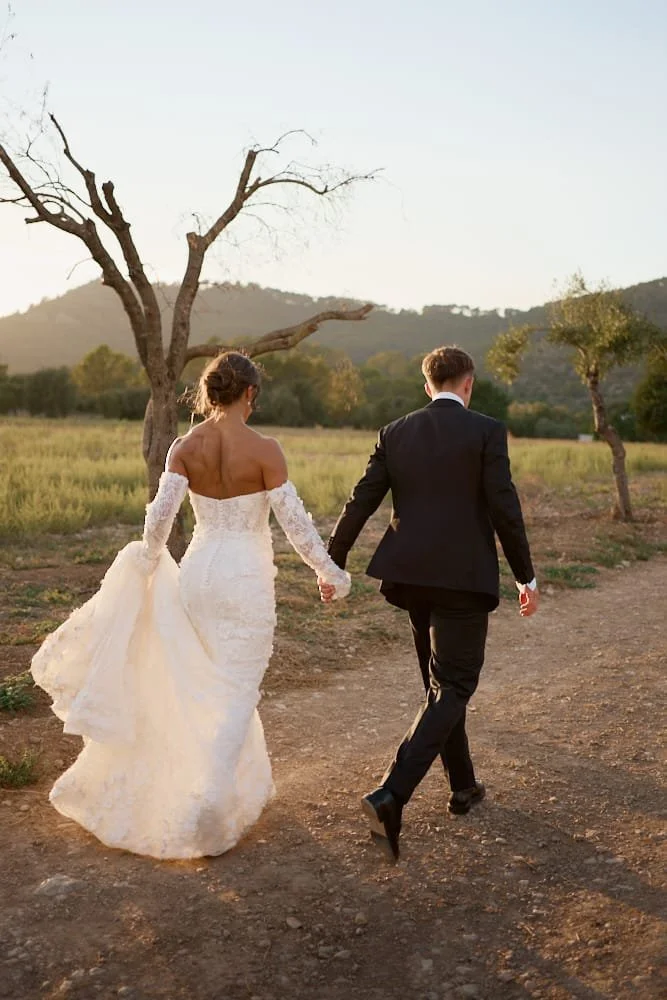 A bride and groom walking hand in hand on a dirt path outdoors during sunset, with trees and hills in the background.