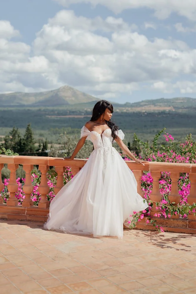 A woman in a white wedding dress stands on a balcony with pink flowers, overlooking a landscape with mountains under a partly cloudy sky.