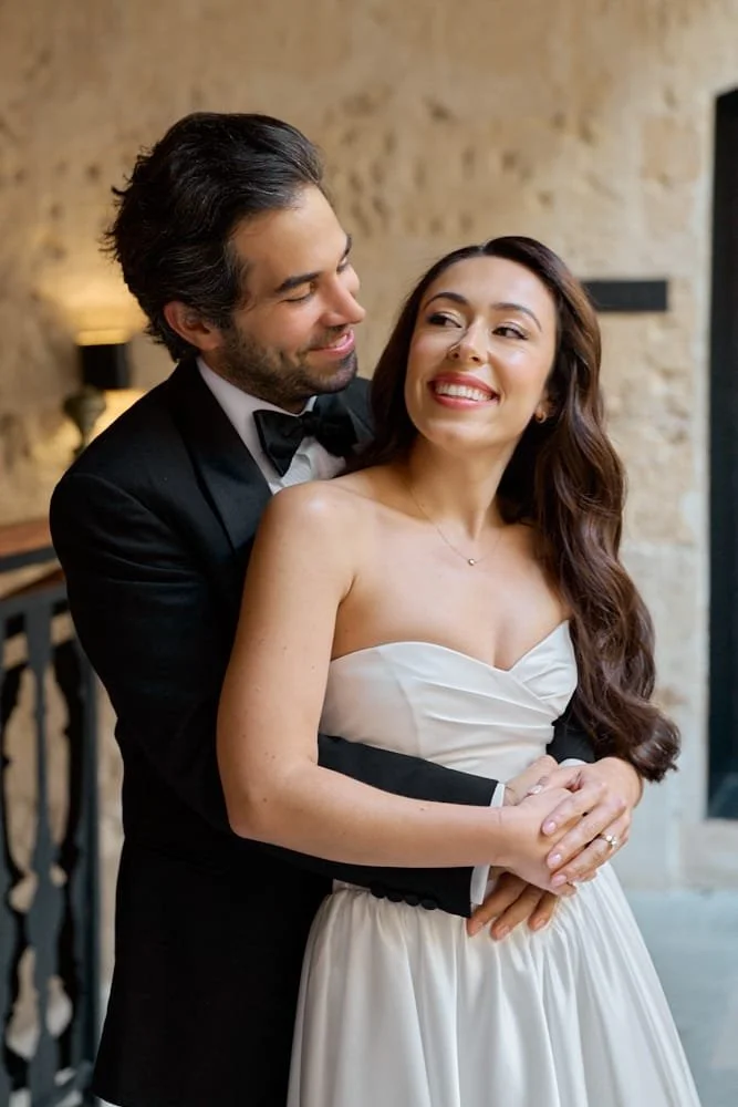 A bride and groom in wedding attire smiling affectionately indoors.