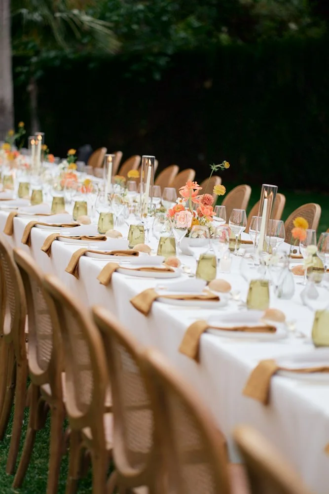 Long outdoor dining table decorated with elegant place settings, glassware, and floral centerpieces, set in a green garden area.