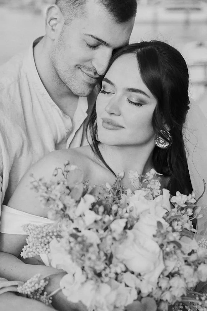 A black and white photo of a man and woman in a close, intimate pose, with the man resting his head on the woman's forehead, she has a bouquet of flowers, and both have their eyes closed.