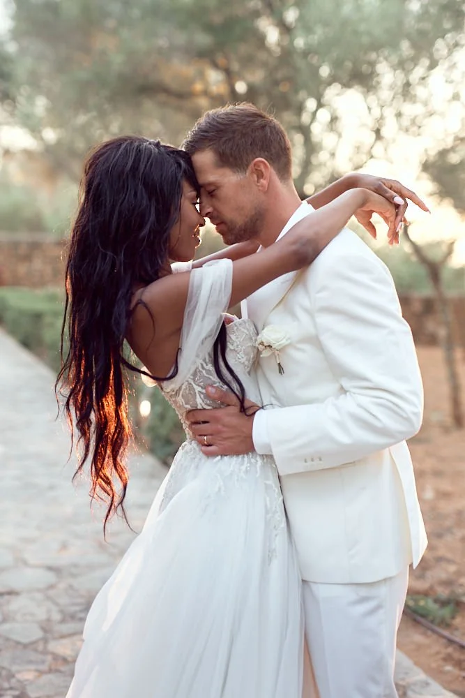 A bride and groom embrace outdoors during sunset, with their foreheads touching, dressed in wedding attire, surrounded by trees and natural scenery.