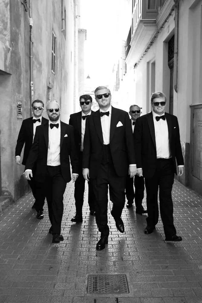 Six men in tuxedos and sunglasses walking down a narrow city alleyway.