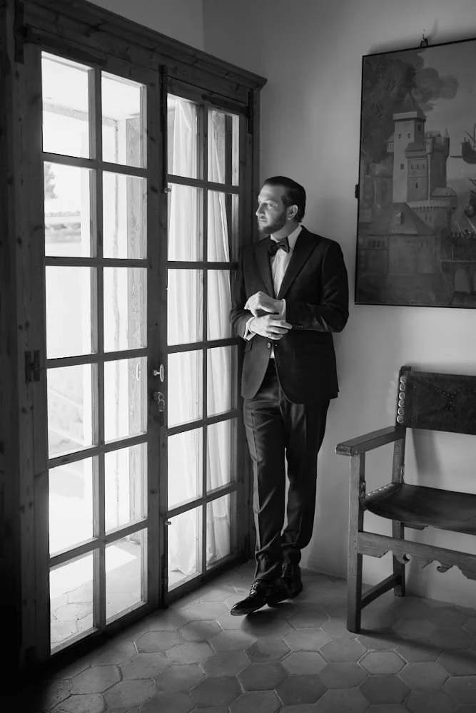 A man in a tuxedo with a bowtie standing by a glass door, looking out thoughtfully with his hands clasped. There is a painting of a castle hanging on the wall and a wooden bench against the wall.