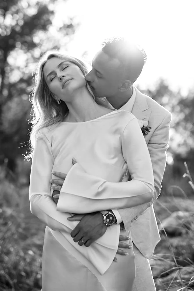 A black-and-white photo of a couple embracing outdoors, with the man kissing the woman's neck. The woman has her eyes closed and appears to be enjoying the moment, while the man is wearing a light suit and a watch. The background shows trees and sunl