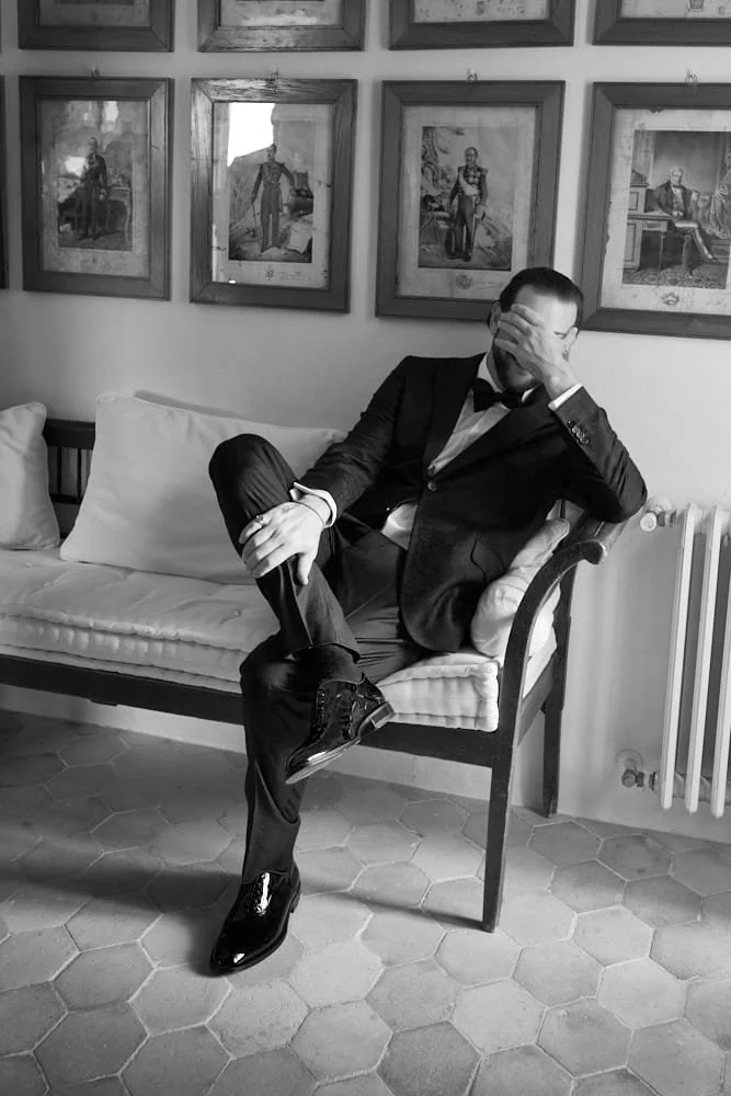A man in a tuxedo sitting on a bench, covering his face with his hand, with framed portraits on the wall behind him.