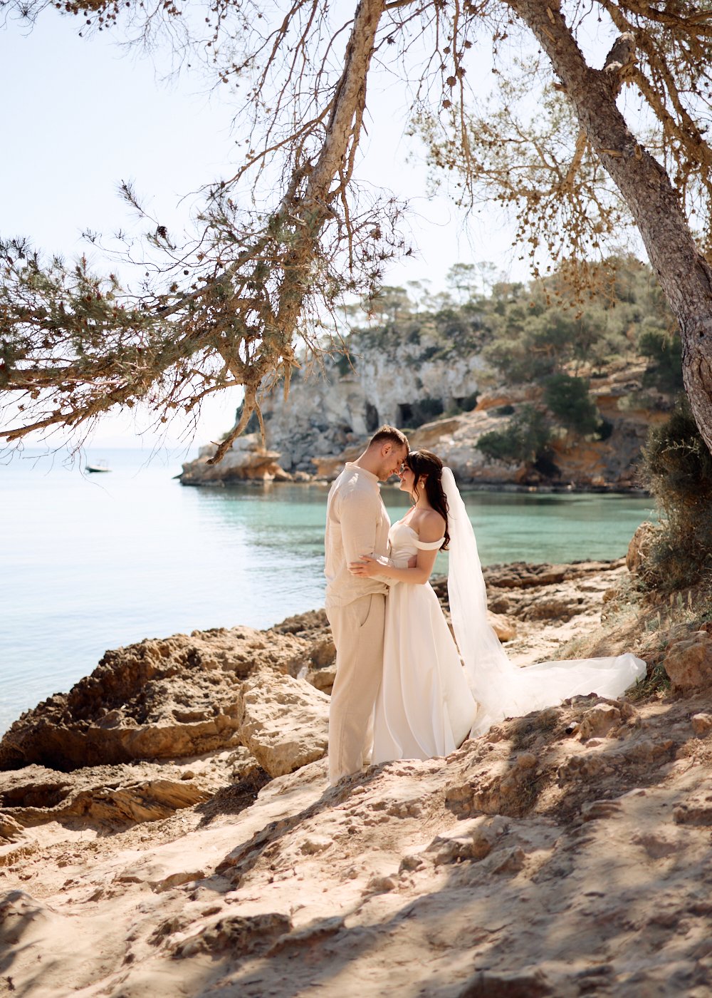 A couple in wedding attire embracing on a rocky beach under a tree, with a calm sea and cliffs in the background.