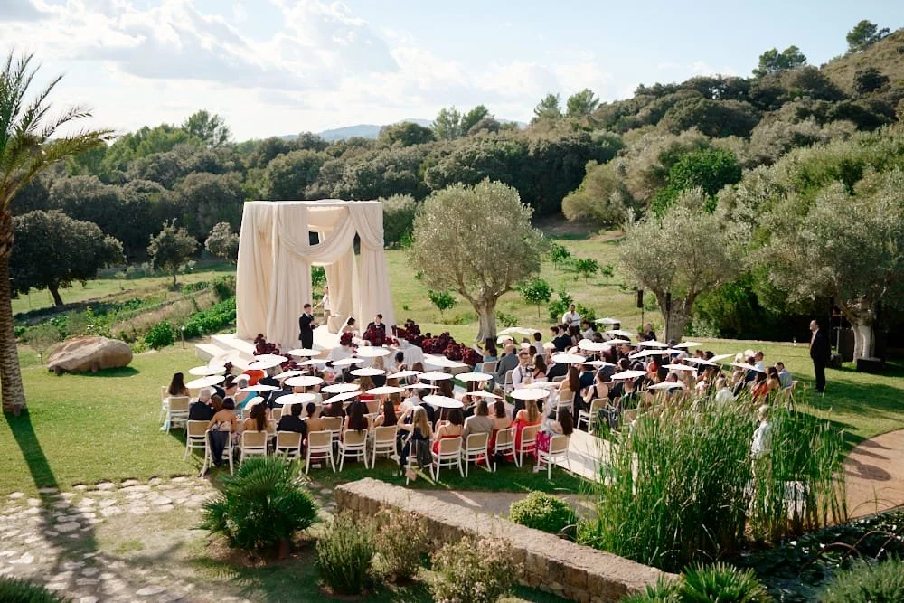 Outdoor wedding ceremony setup with white chairs, a draped arch and many guests with umbrellas, surrounded by trees and greenery.