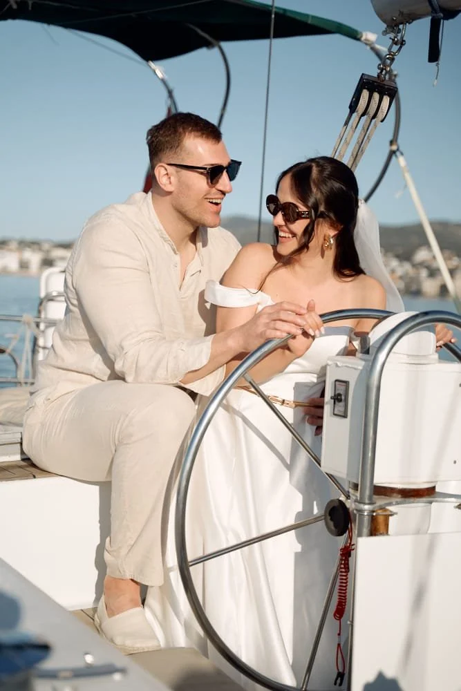 A happy couple dressed in wedding attire on a sailboat, smiling and holding hands with the woman in a white dress and sunglasses, the man in light-colored clothing and sunglasses, with a background of water and a distant shoreline.