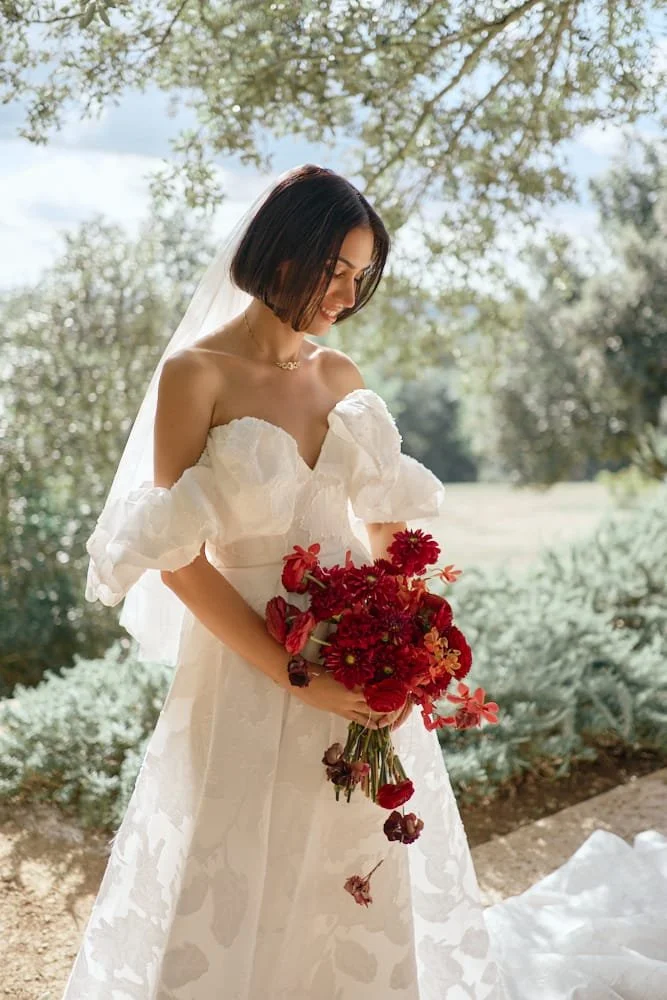 A bride in a white off-shoulder wedding dress standing outdoors during daytime, holding a bouquet of red flowers, with trees and a bright natural background.