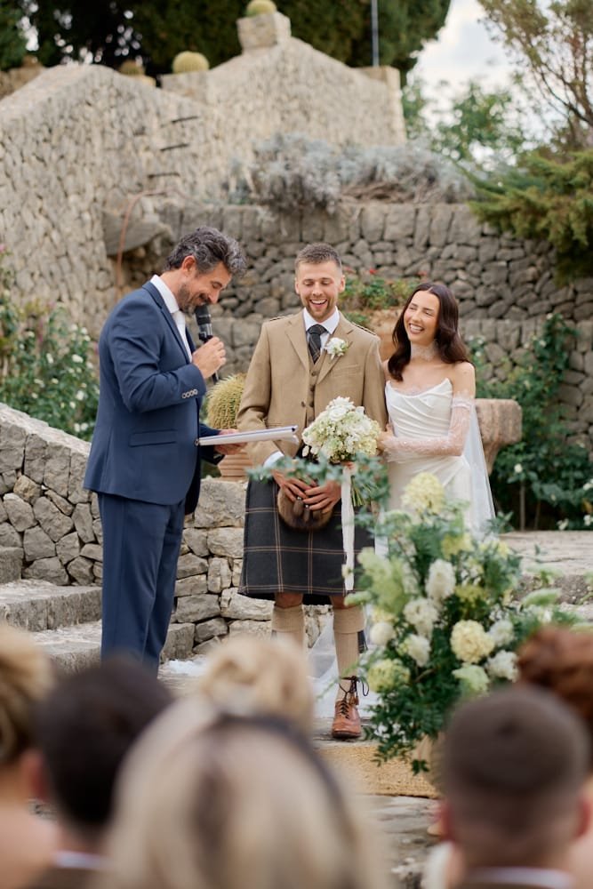 A wedding ceremony outdoors with a couple and officiant, surrounded by flowers and stone structures.