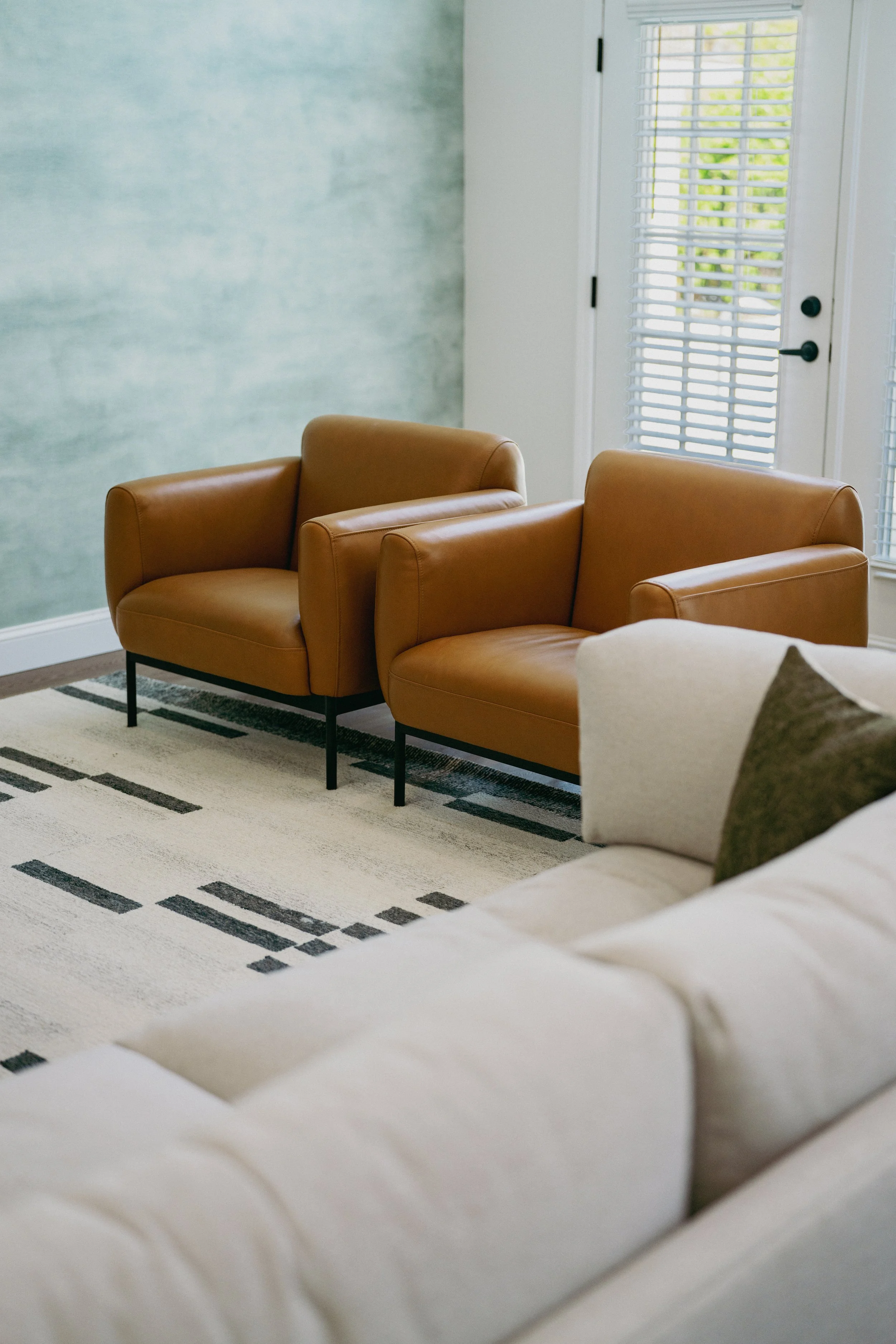 Living room with two tan leather armchairs against a light blue wall, large window with white blinds, and a cream sofa with a green pillow in the foreground.