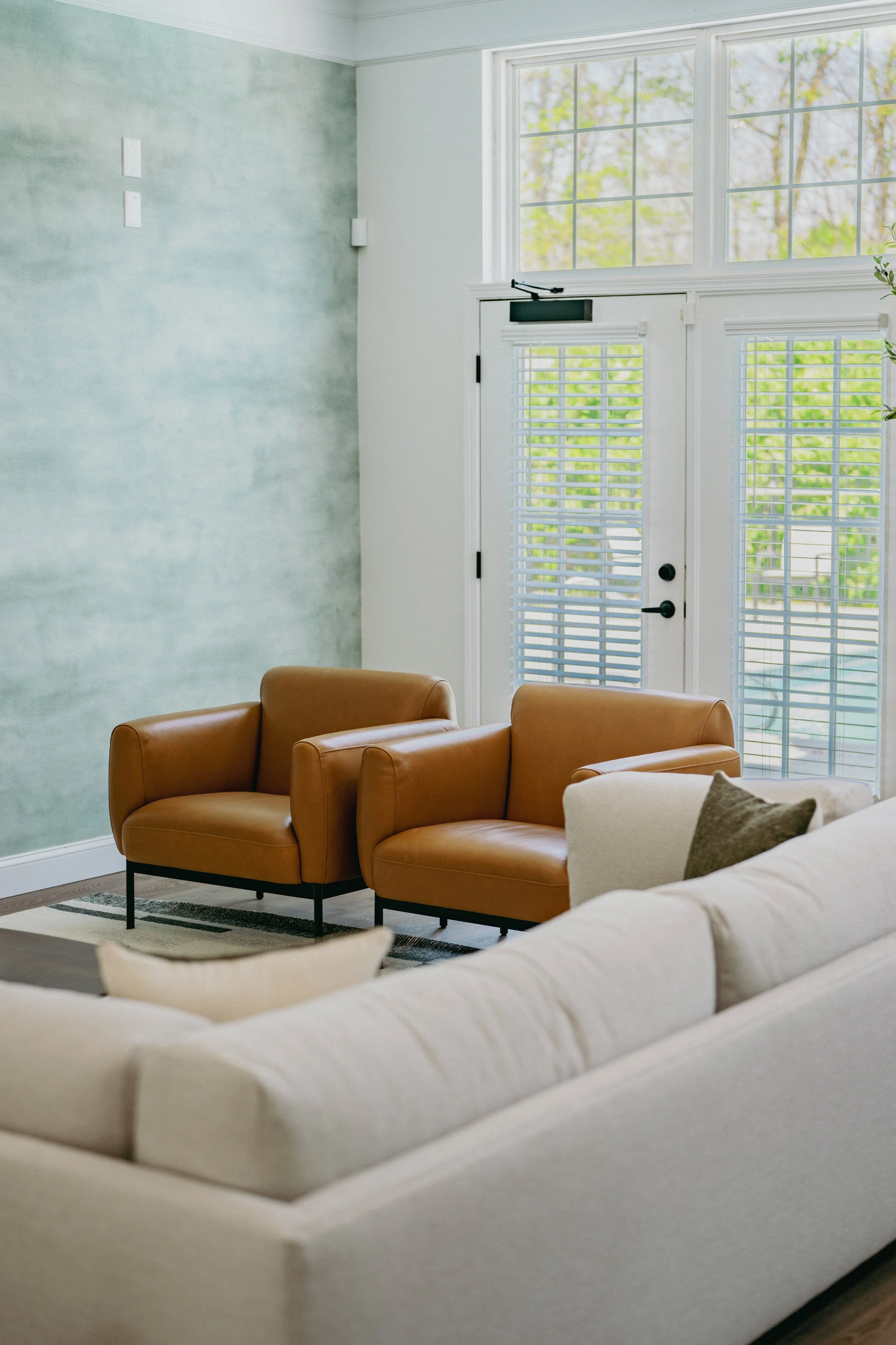 A living room with two tan leather armchairs, a white sofa, and a large window with white blinds, overlooking trees outside.