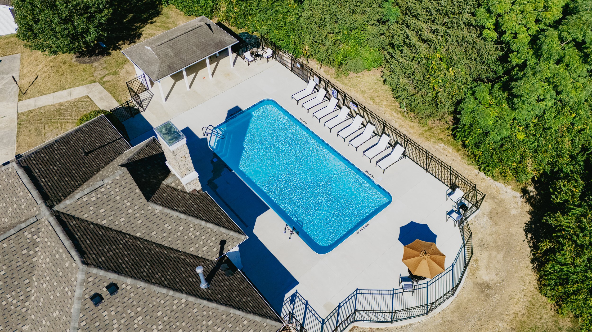 Aerial view of a backyard swimming pool with white lounge chairs around it, a small covered patio area, and green trees and bushes surrounding the yard.