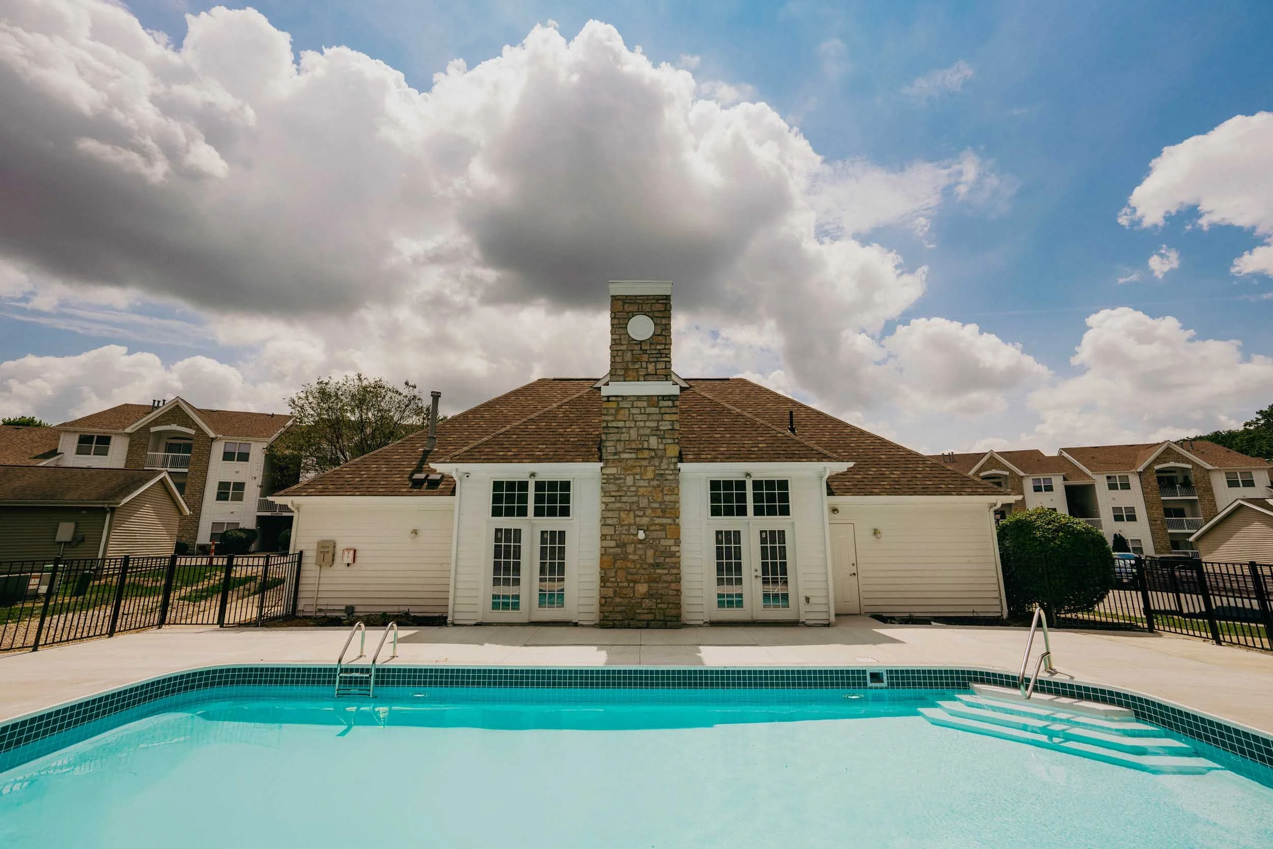 View of a backyard with a swimming pool in the foreground, white house with a stone chimney and multiple windows behind, and a cloudy sky overhead.
