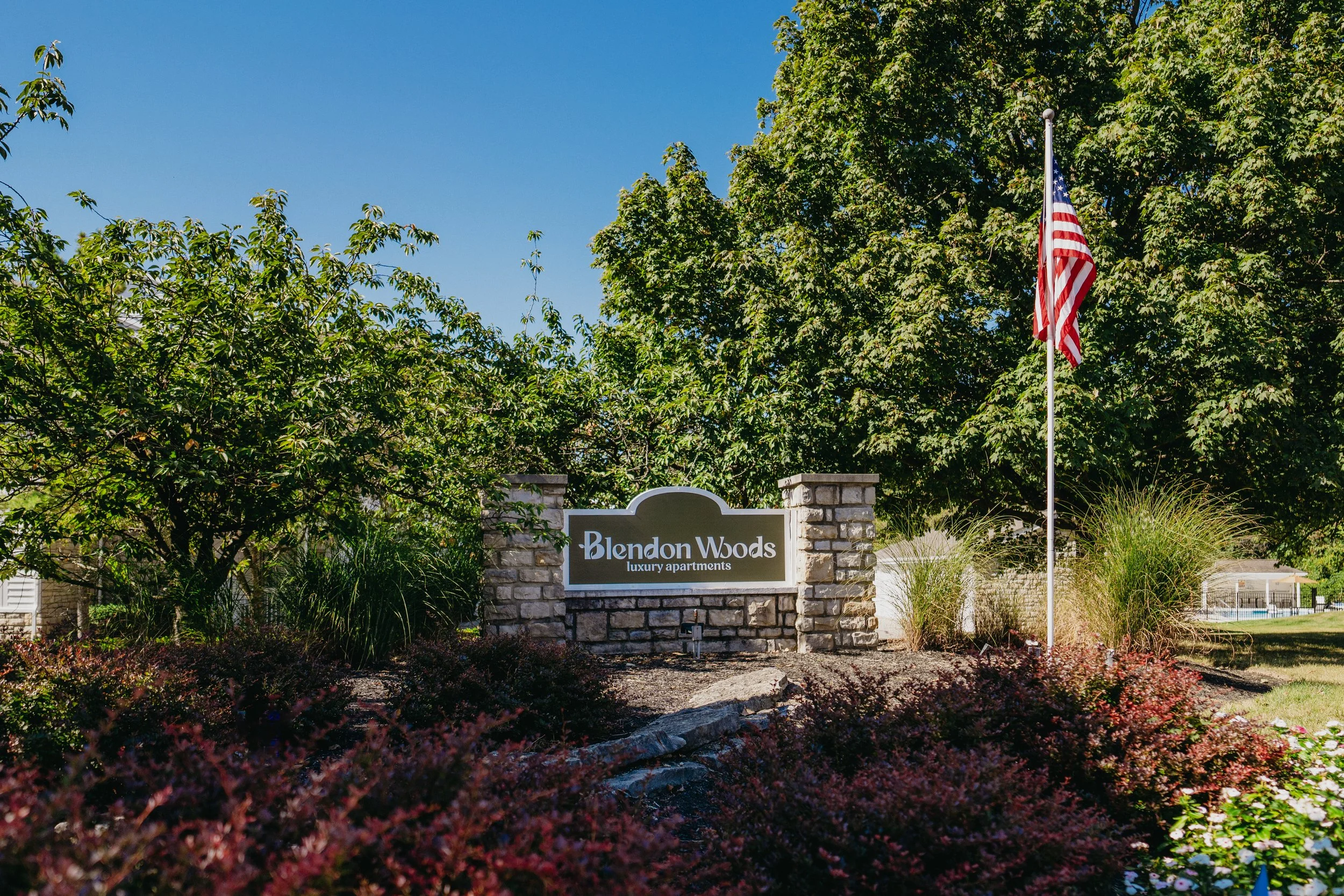 Entrance sign for Blendon Woods luxury apartments with landscaping, trees, and an American flag on a flagpole under a clear blue sky.
