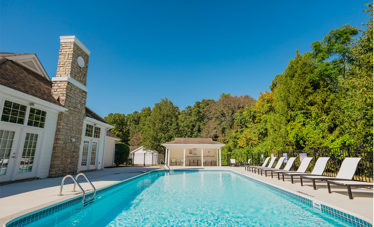 An outdoor swimming pool with lounge chairs along the fence, a small pool house, a house with a stone chimney, and trees in the background under a clear blue sky.