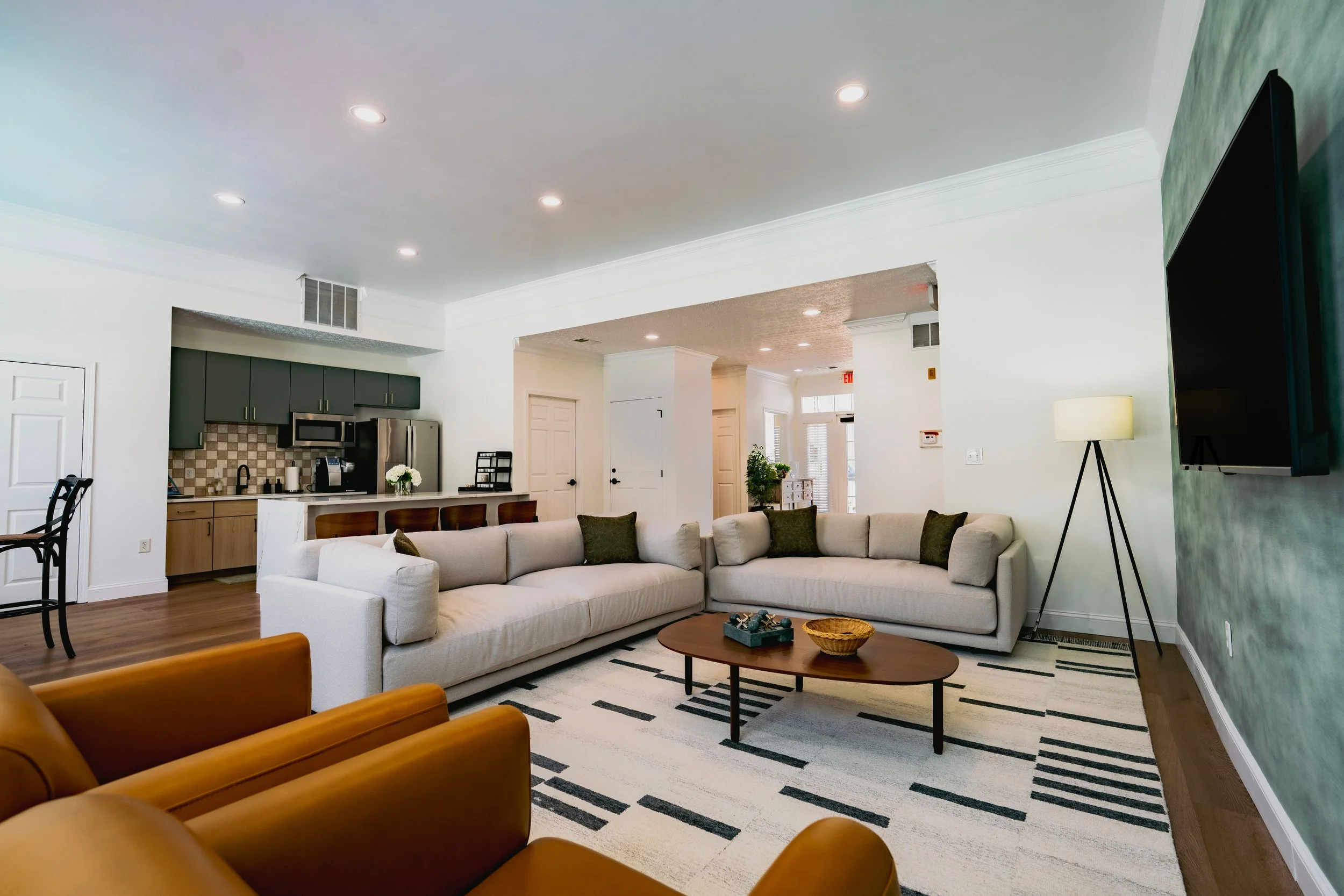 Living room with white sectional sofa, black and white striped rug, wooden coffee table, green accent wall, flat-screen TV, standing lamp, and part of a dining area with chairs and kitchen in the background.