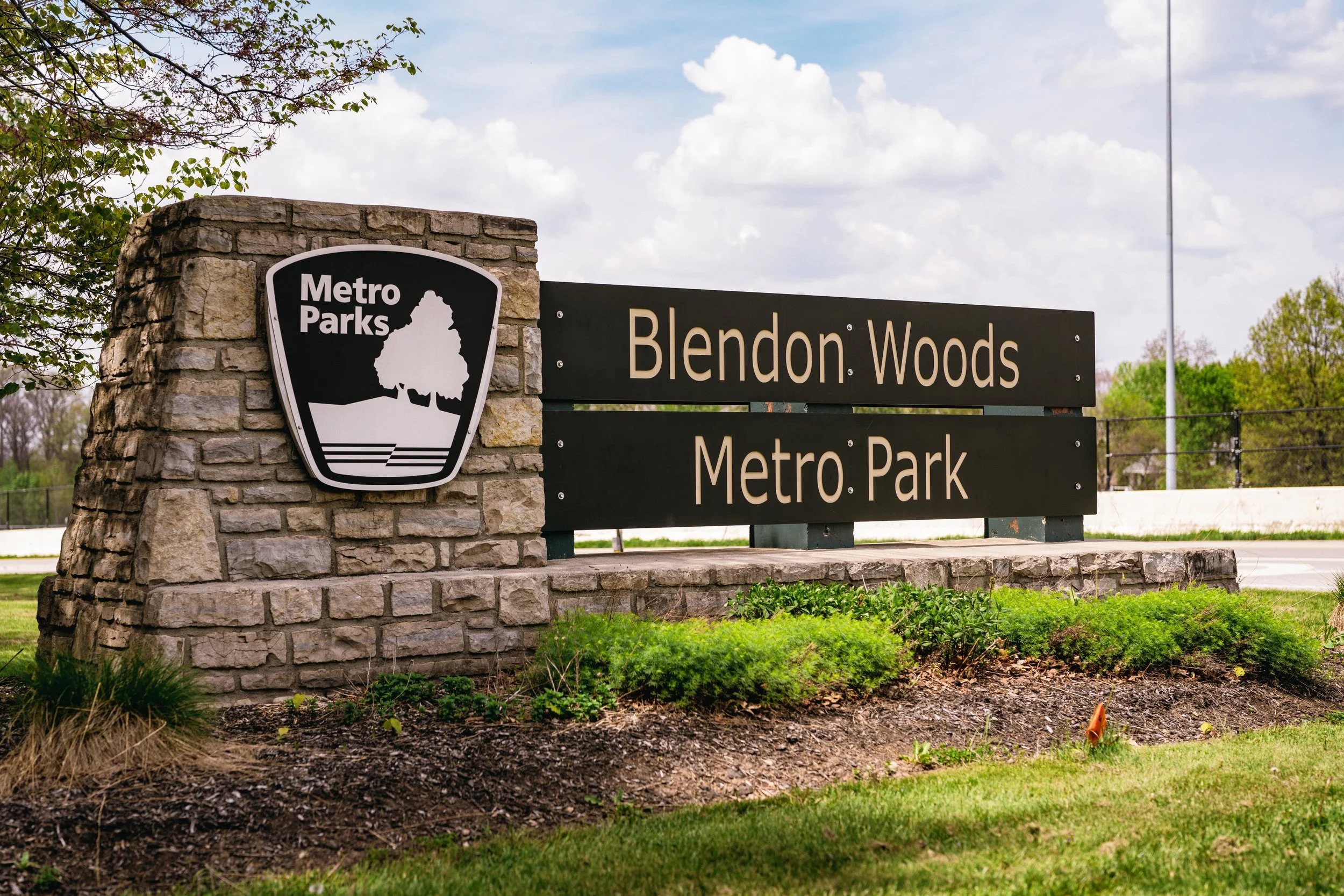 Sign at the entrance of Blendon Woods Metro Park with the Metro Parks logo, featuring a tree silhouette, mounted on a stone pillar, surrounded by greenery and a partly cloudy sky.