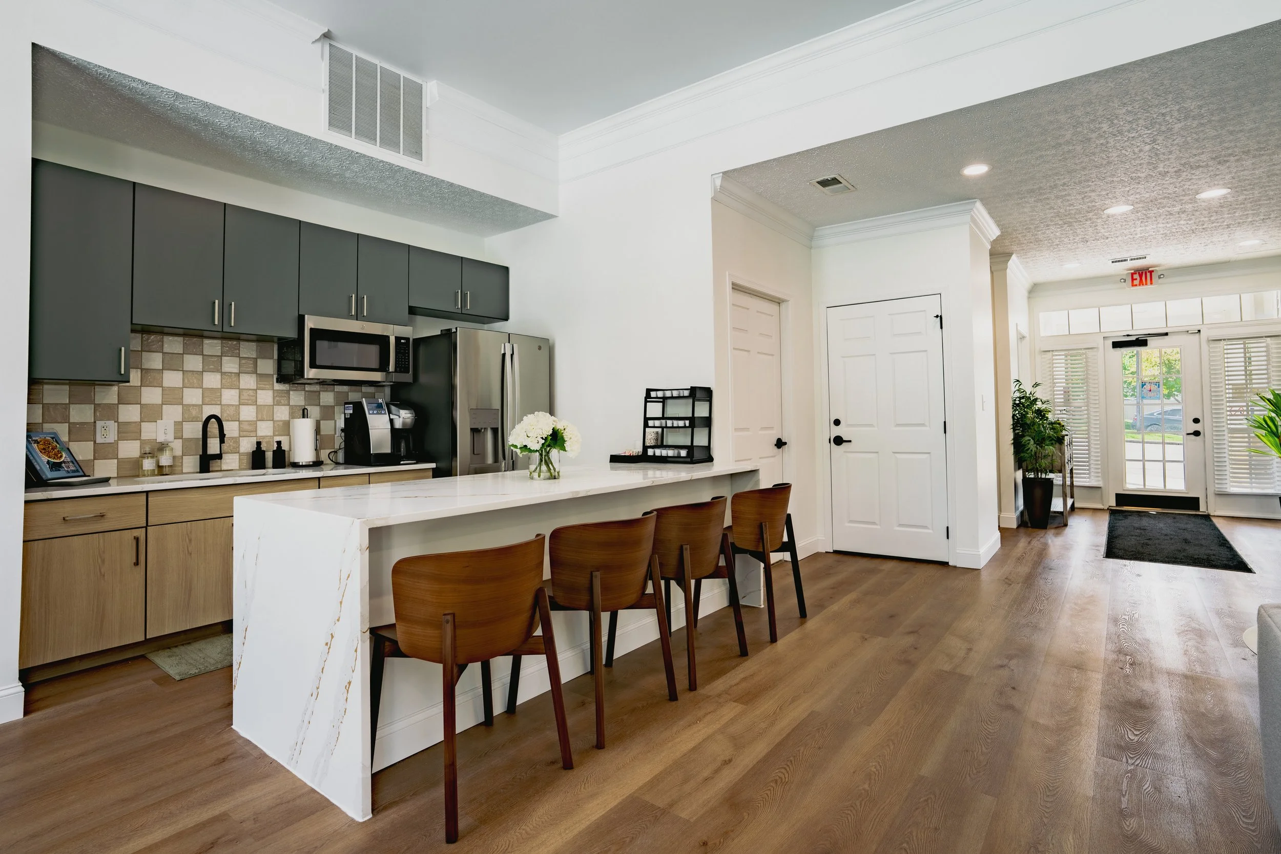 Modern kitchen with gray cabinets, stainless steel appliances, a white marble island, and a dining area with wooden chairs near the entrance with glass doors and white blinds.