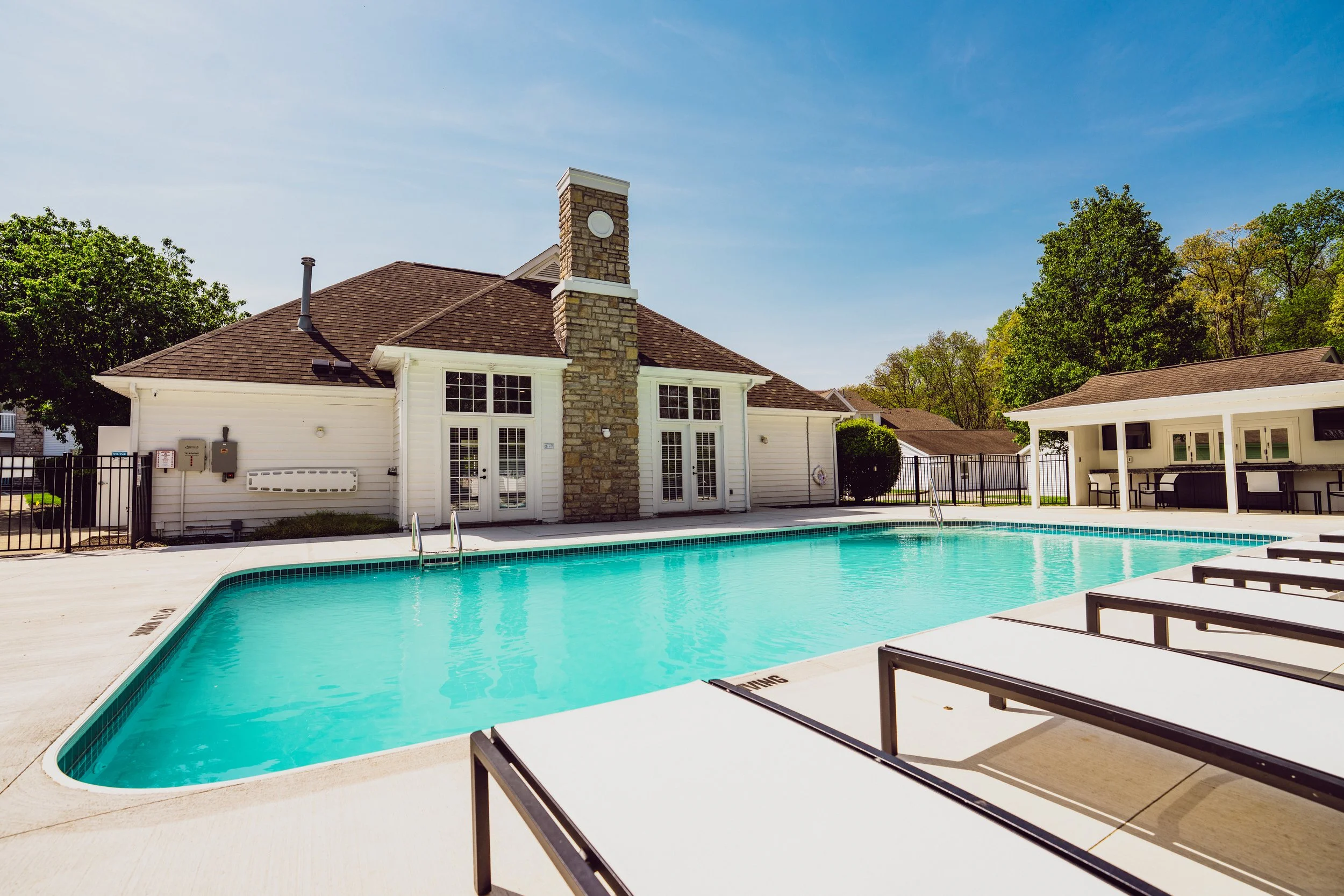 A backyard swimming pool with lounge chairs, surrounded by a black fence, with a house featuring a stone chimney and a patio area in the background. Clear blue sky and green trees are visible.