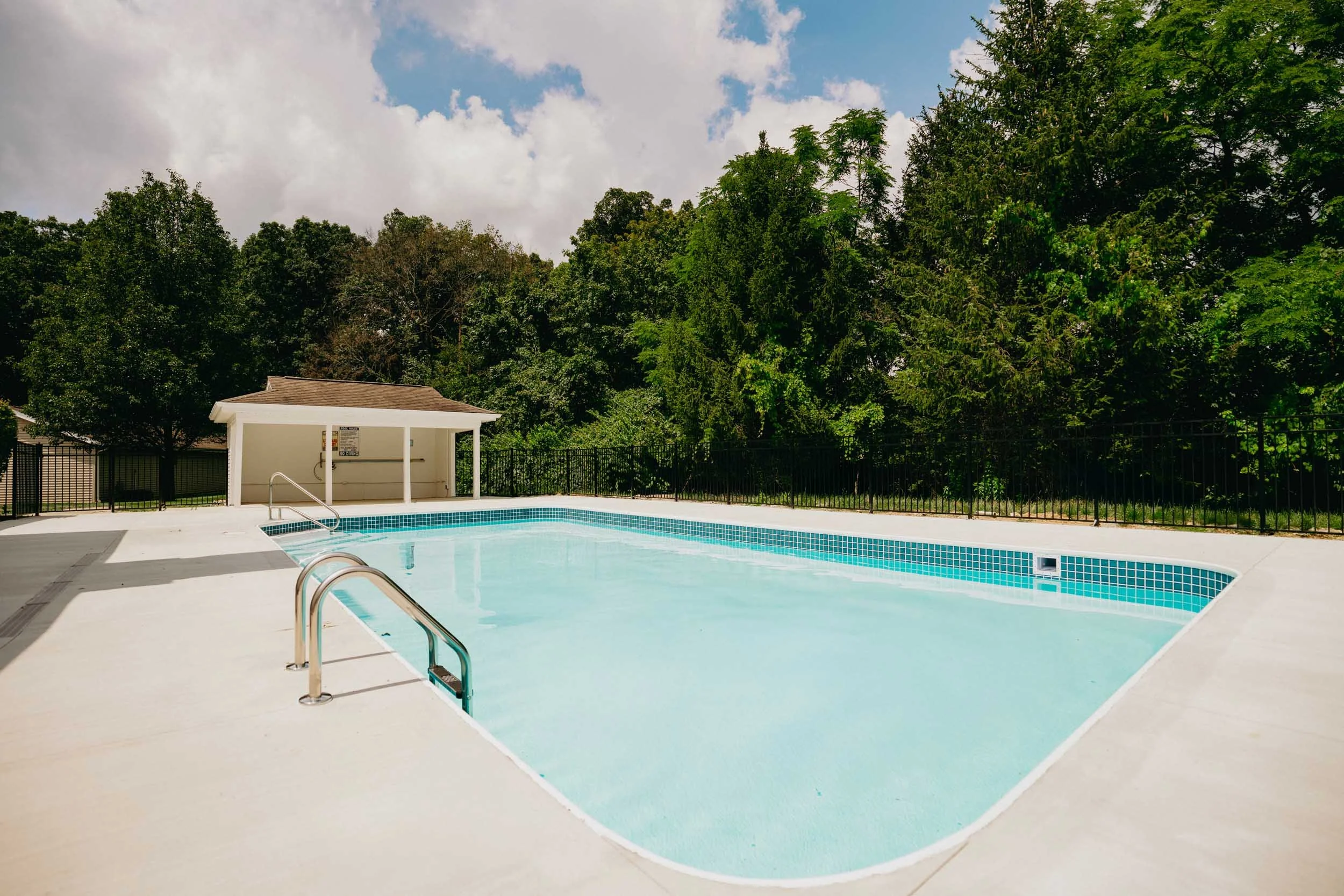 Empty outdoor swimming pool with blue tile accents, black fence, and surrounding greenery under partly cloudy sky.