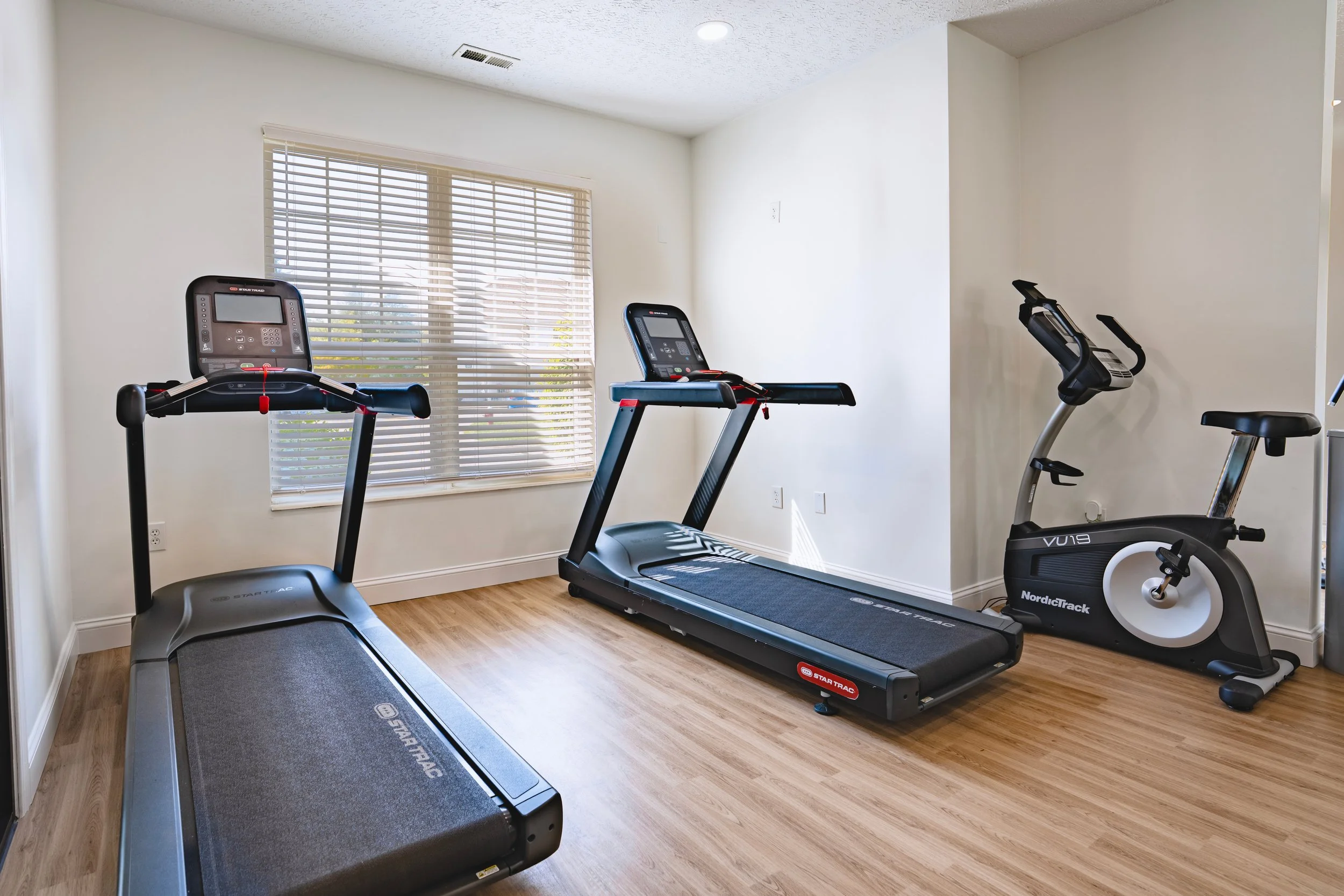 Home gym with treadmill and stationary bike near window with blinds, hardwood floor, white walls, and ceiling light.