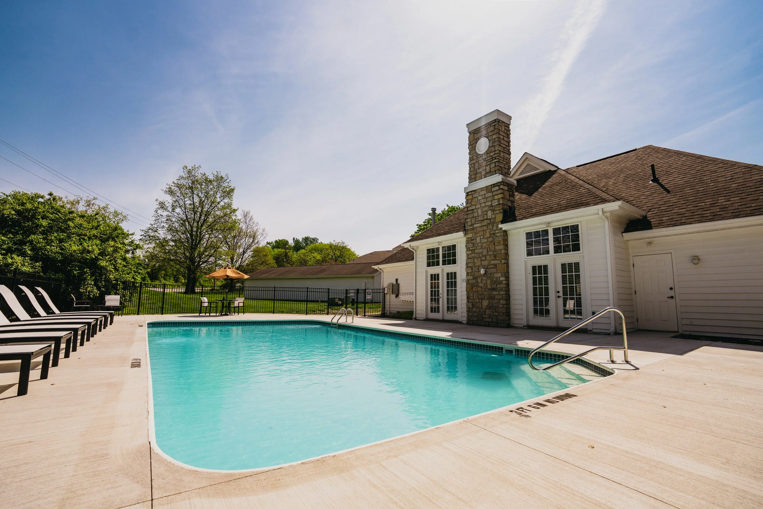 Outdoor swimming pool with lounge chairs, surrounded by a black fence, with a house featuring a brick chimney and a gable roof in the background, under a clear blue sky.