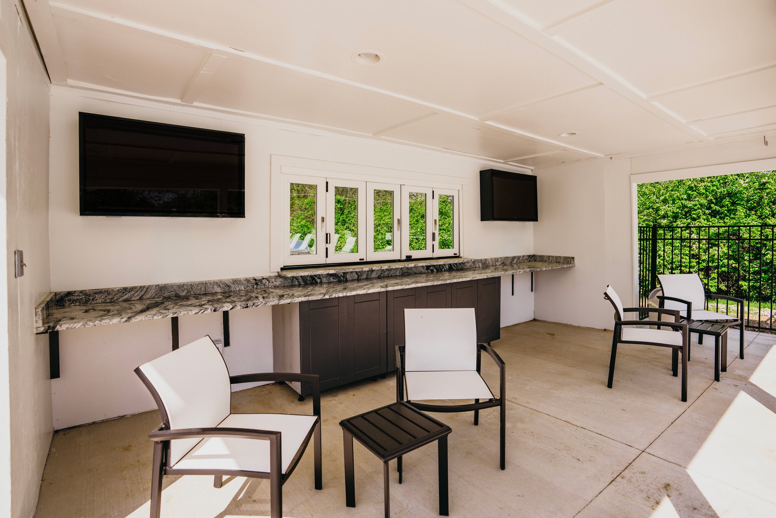 Outdoor porch with white ceiling, two wall-mounted televisions, a marble countertop, black cabinets, white chairs, and green trees outside.