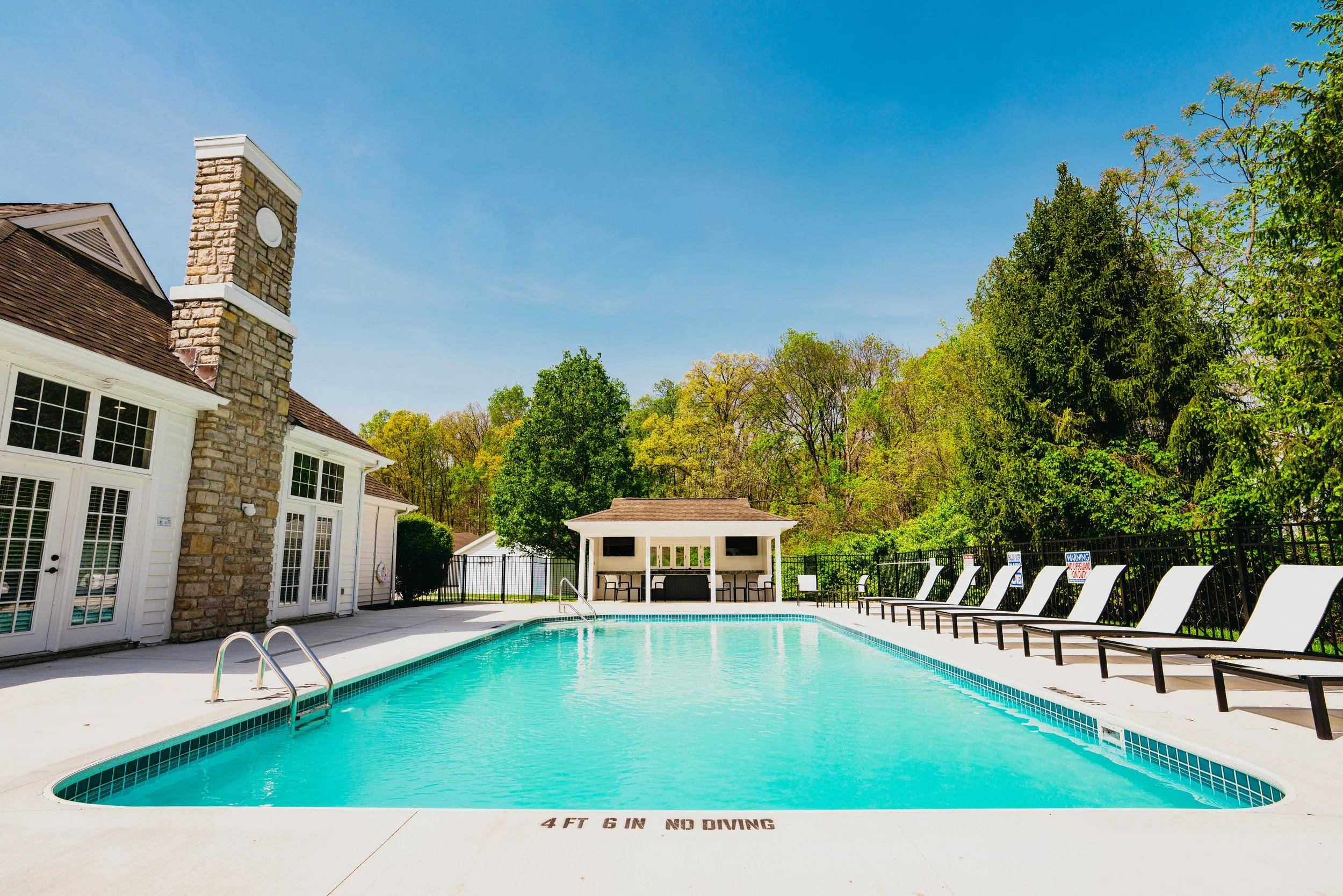 A backyard swimming pool area adjacent to a house with a stone chimney, surrounded by trees under a clear blue sky. The pool has a sign indicating no diving and a row of lounge chairs along the fence.