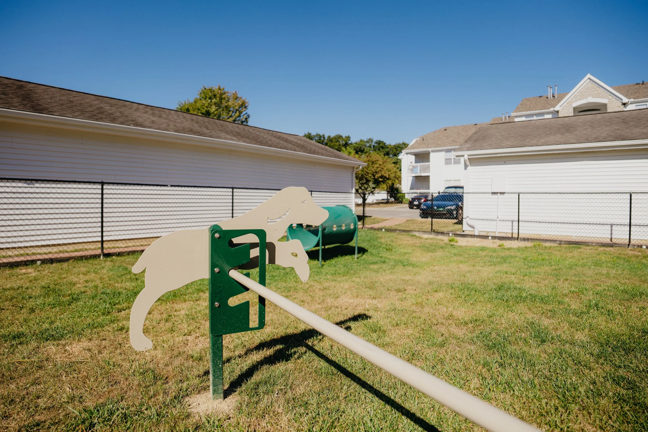 Playground area with a white and green horse-shaped rocking toy on a grassy lawn, surrounded by residential houses and a black chain-link fence under a clear blue sky.
