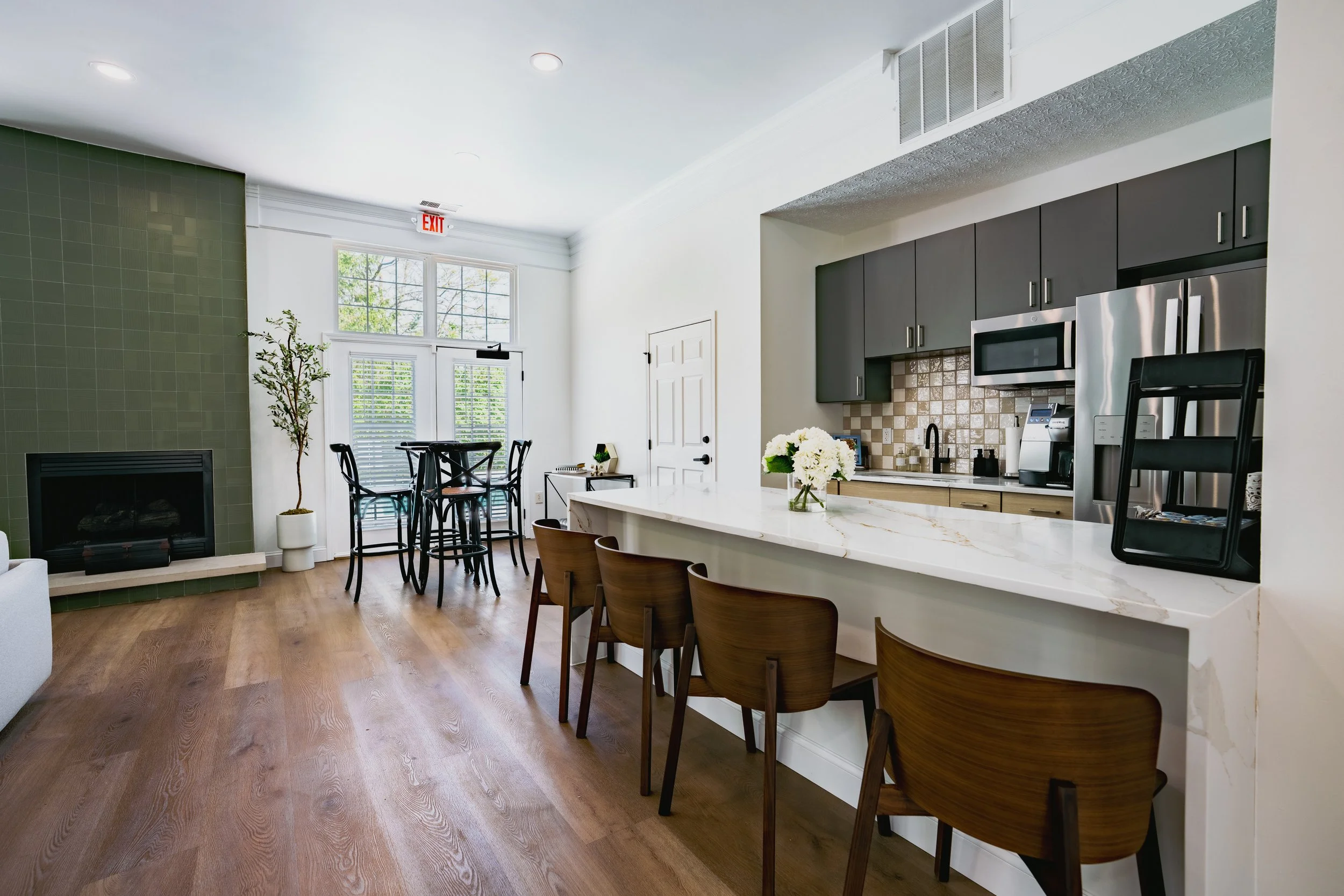 Open plan living area with kitchen, bar stools, dining table, and fireplace, featuring natural light from large windows, modern dark cabinets, and a wood floor.