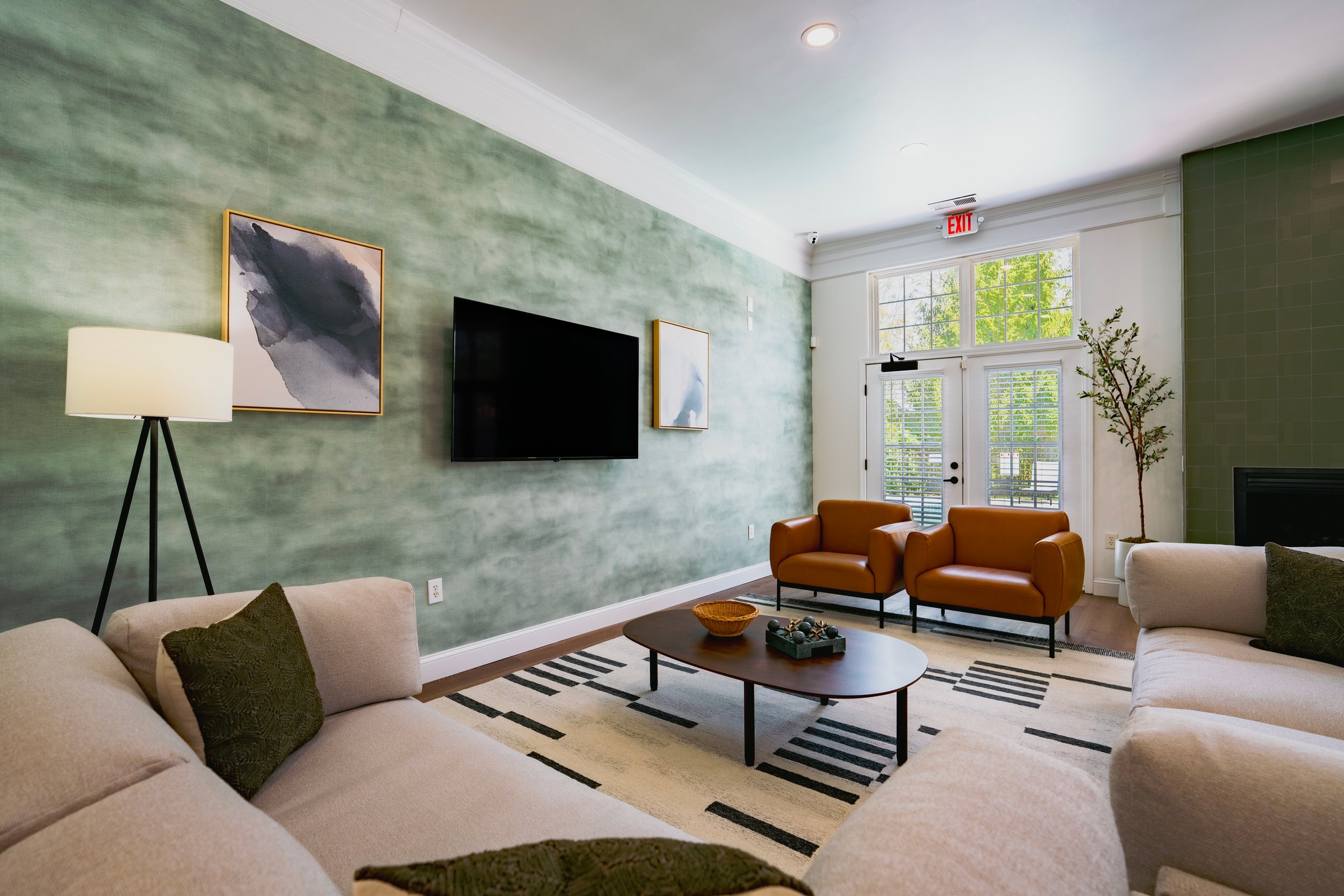 Living room with green accent wall, abstract artwork, wall-mounted TV, beige sofa, orange armchairs, coffee table, striped rug, potted plant, and large windows letting in natural light.