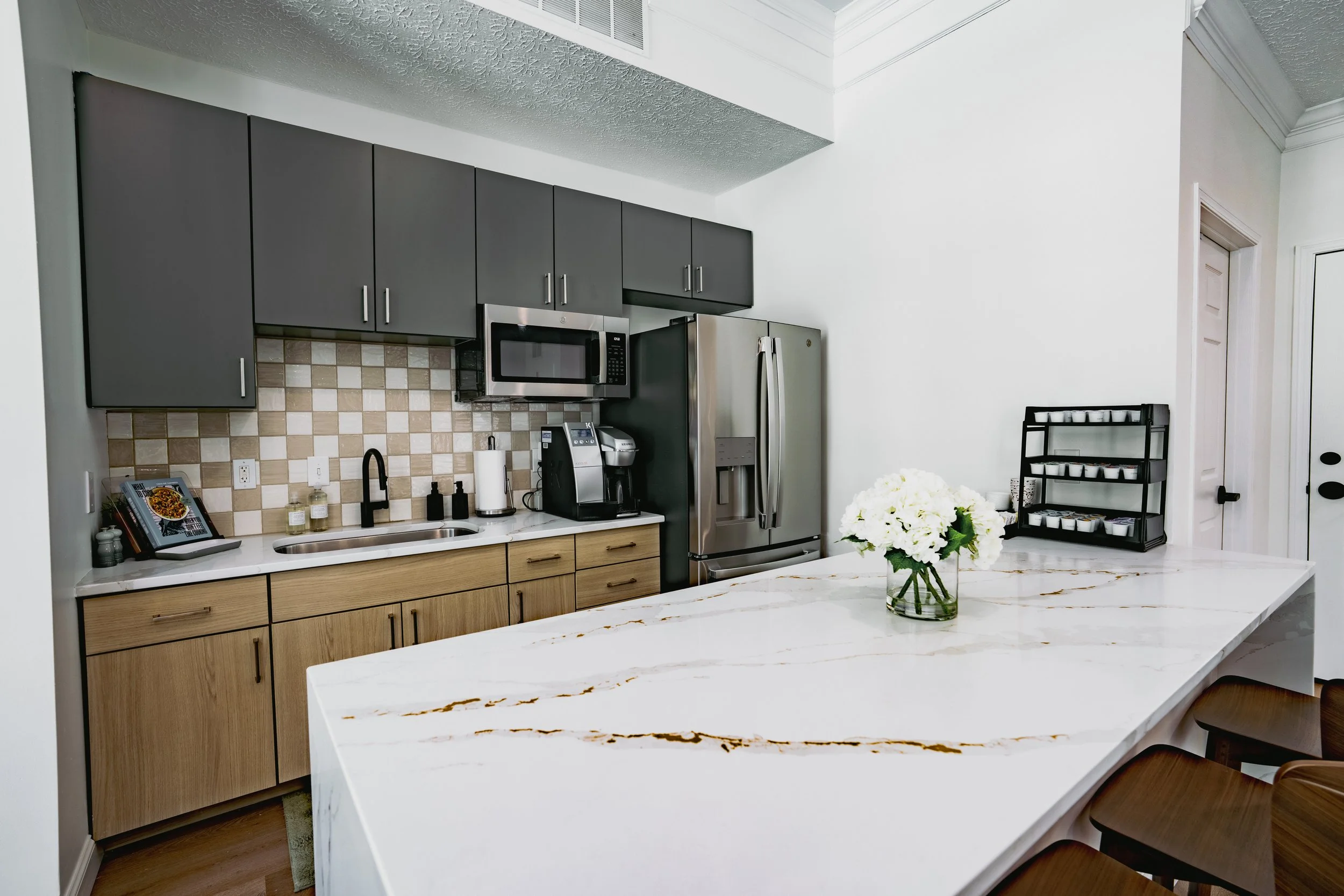 Modern kitchen with gray cabinets, stainless steel refrigerator, microwave, and coffee machine. White marble island with gold veining, a vase of white flowers, and a black dish rack with small white bowls.
