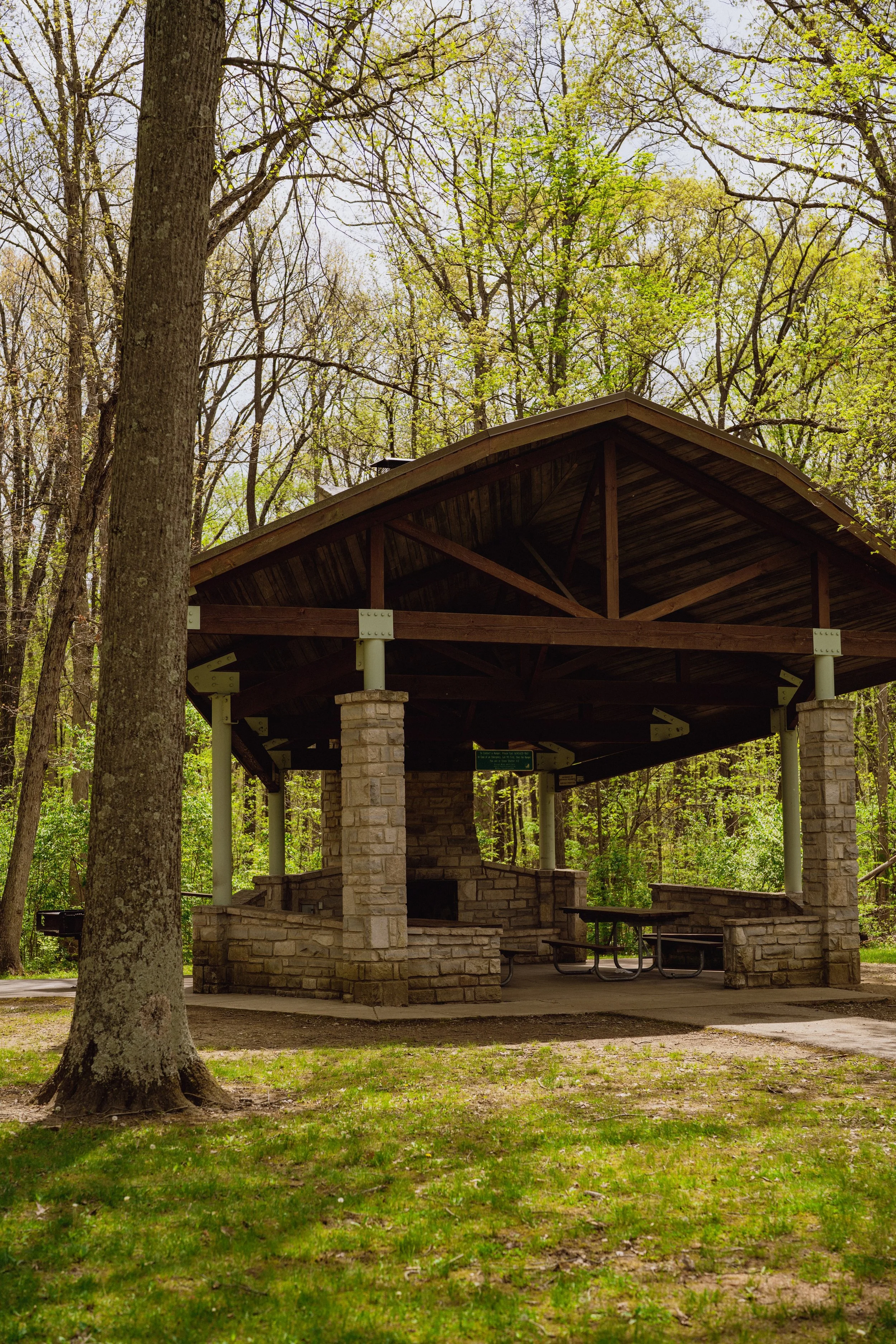 A wooden pavilion with a sloped roof, supported by stone and white metal columns, situated in a wooded park with trees and green grass.