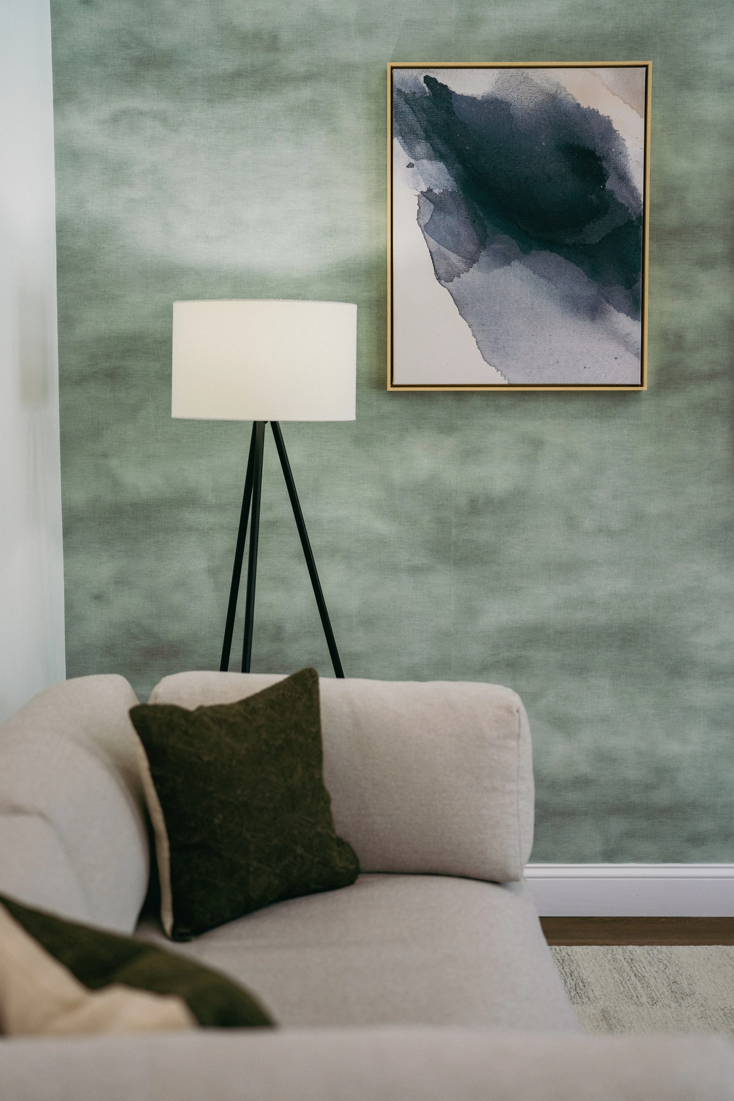 A living room with a white couch, dark green pillow, green textured wall, framed abstract art, and a white floor lamp with black legs.