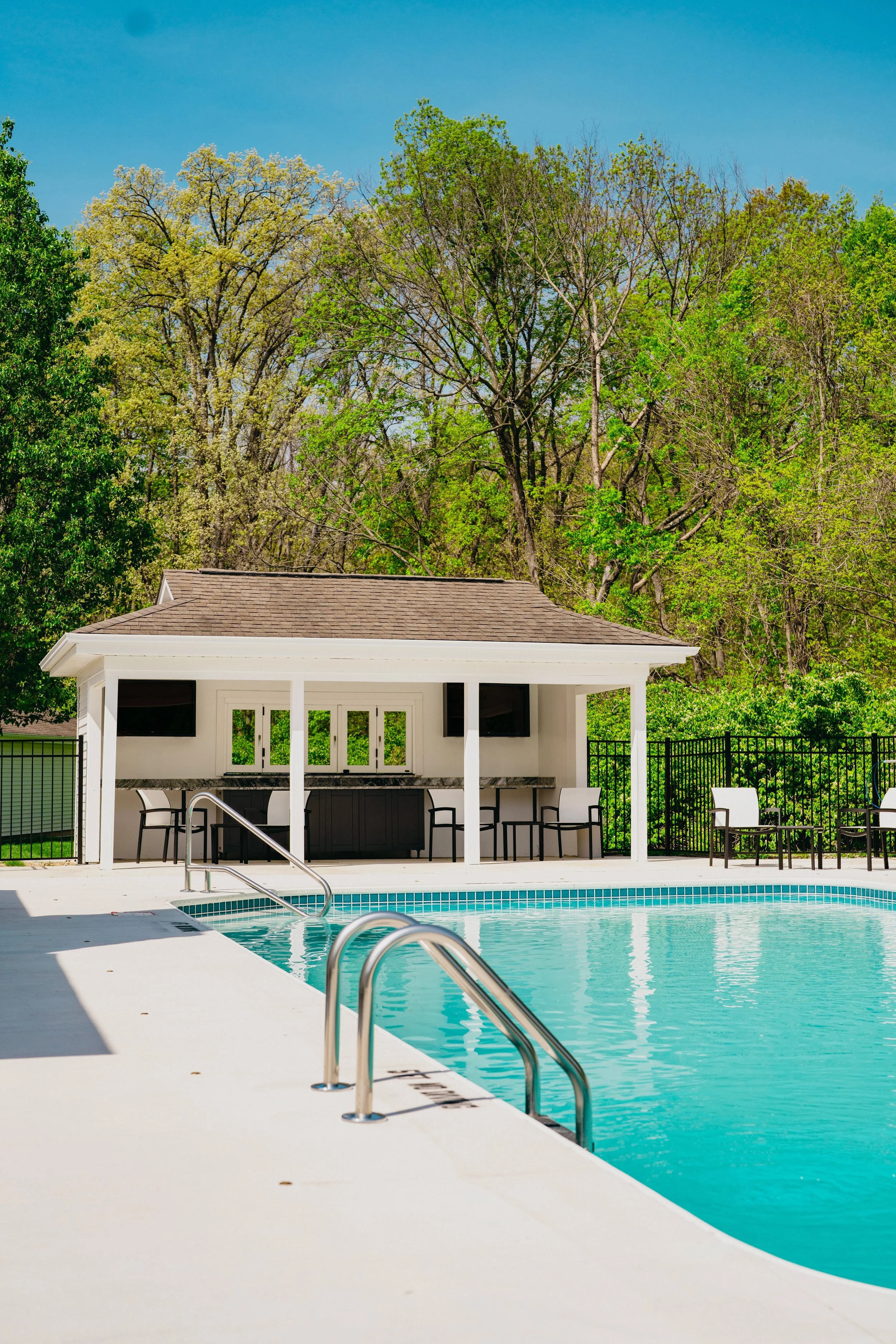 Empty swimming pool with metal handrails, surrounded by a white pool deck and a covered lounge area with chairs and bar, set against green trees and a clear blue sky.